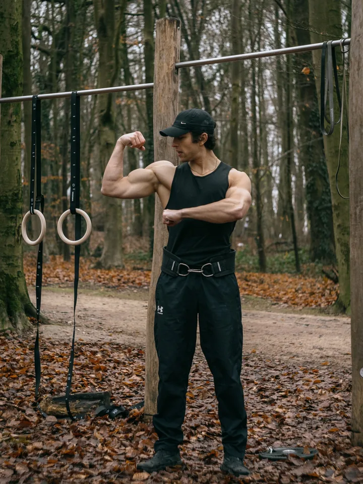 Marius Grek, casquette et débardeur noirs, ceinture de lest à la taille, devant des barres de traction et des anneaux de gymnastique, dans un bois en automne